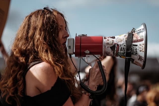 A imagem mostra uma jovem mulher segurando um megafone em frente a sua boca. Ela está de roupa preta e o megafone é branco e vermelho com alguns adesivos coloridos.
