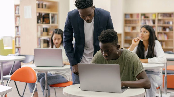 Students learning in a classroom setting with a teacher assisting and laptops on desks, creating an interactive education environment.